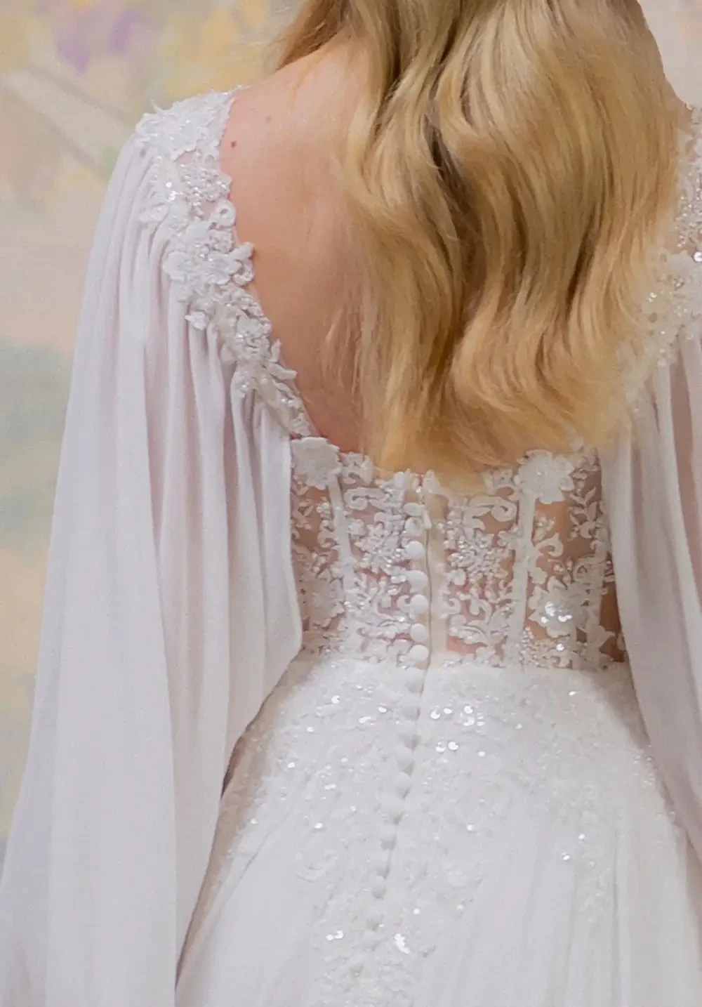 A close-up view of the back of a wedding dress featuring intricate lace detailing and buttons, with long, flowing sleeves.