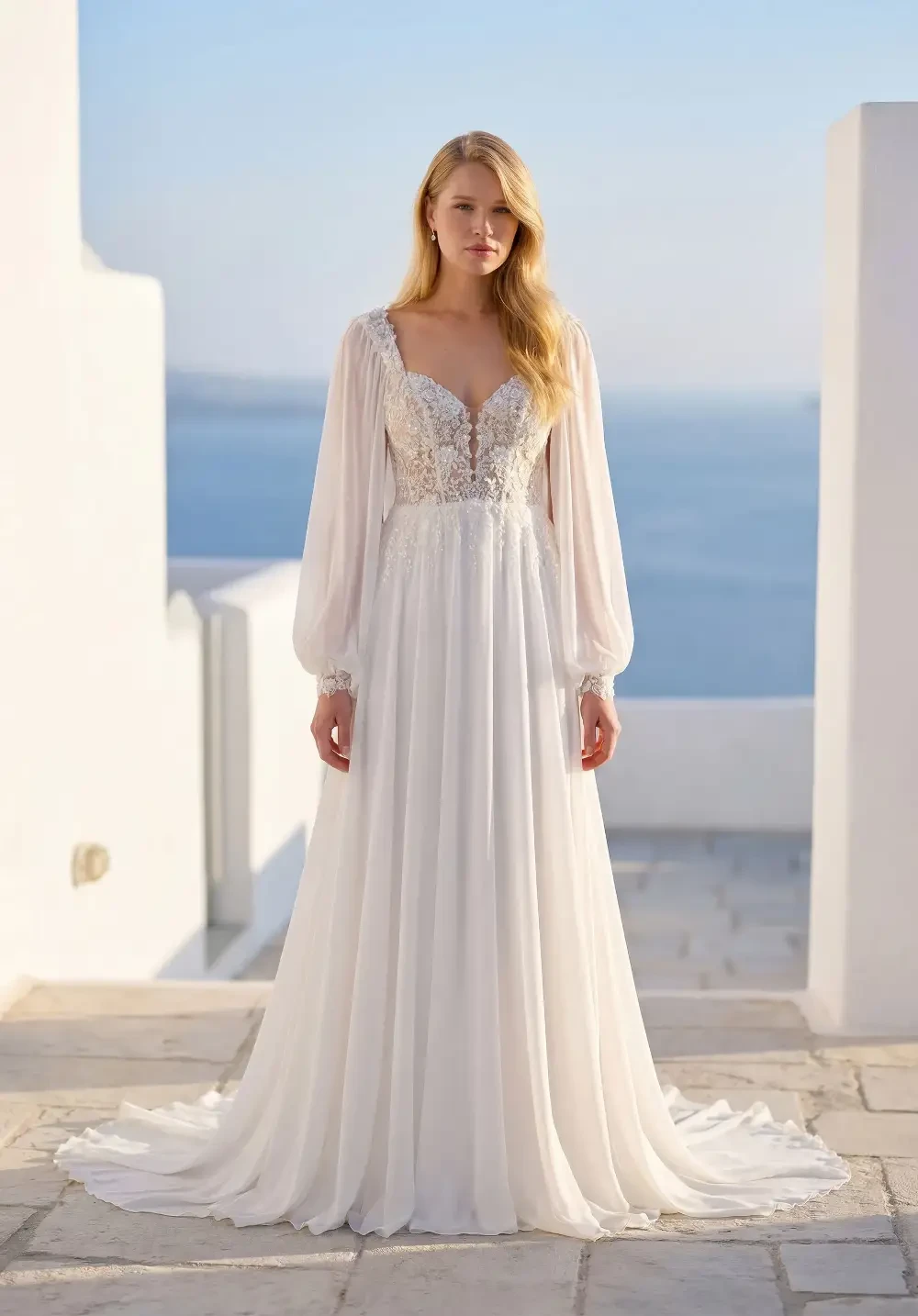 A woman in a flowing white dress with long sleeves and lace detailing stands in front of a serene blue ocean background.