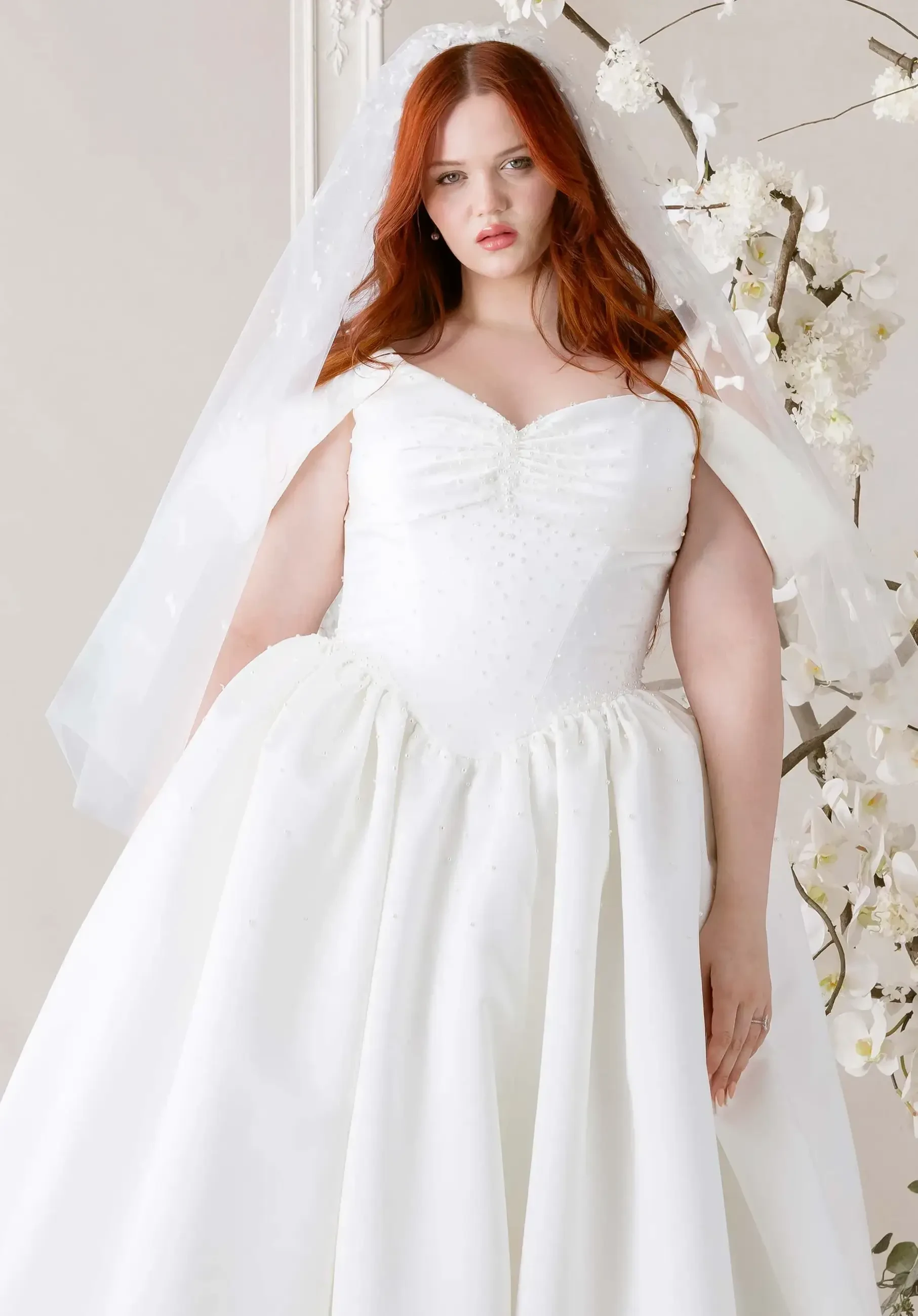 A woman with long red hair is wearing a white wedding dress and veil, standing in front of a floral backdrop.