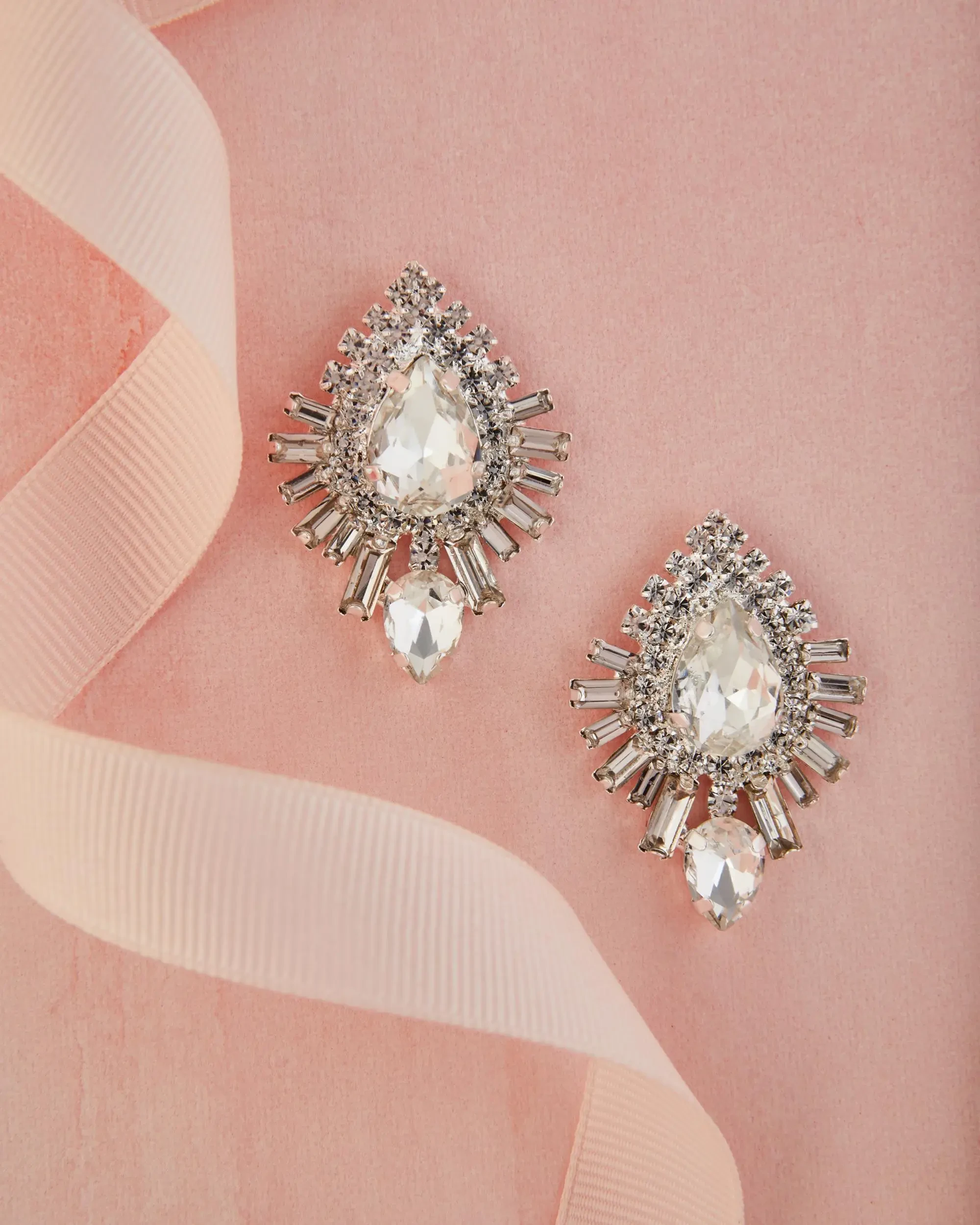 A pair of teardrop-shaped earrings with sparkling clear gemstones set in a silver frame, displayed on a pink background alongside a soft ribbon.