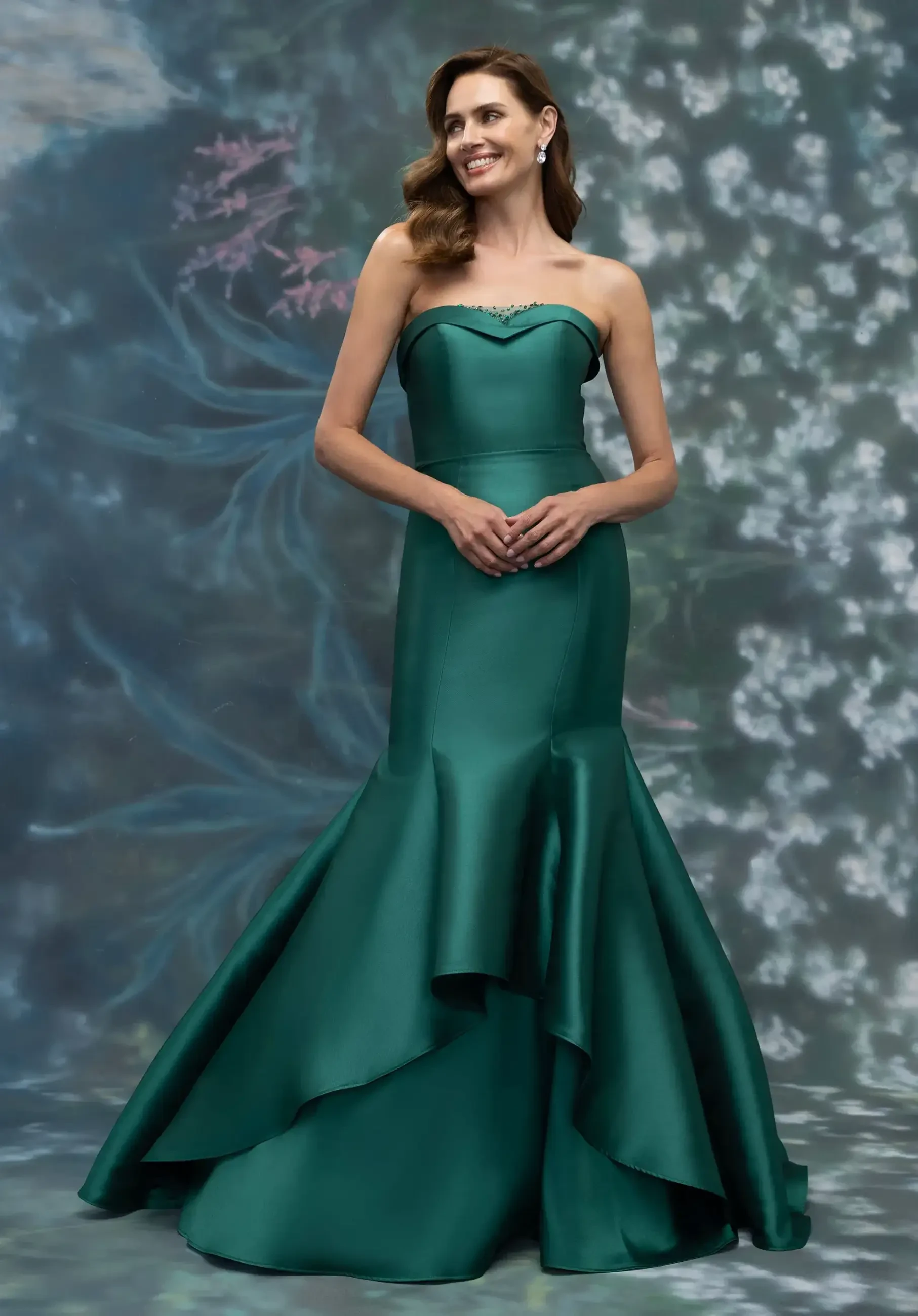A woman in a strapless, emerald green gown with a mermaid silhouette, standing against a floral backdrop.