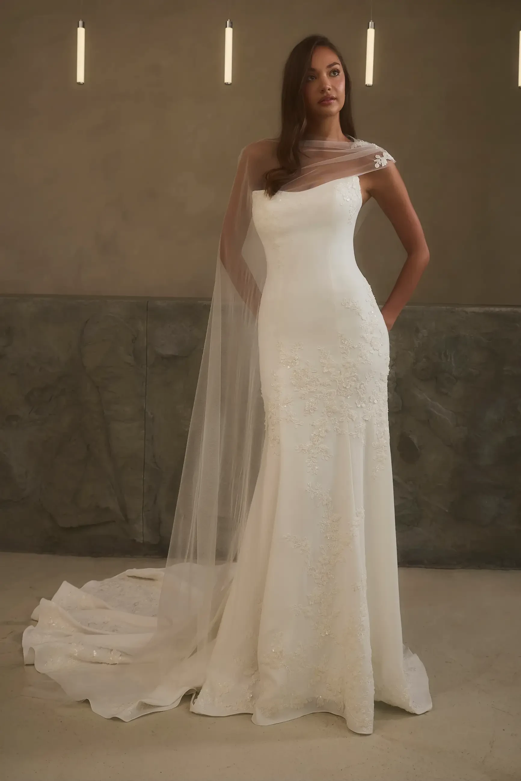 A model wearing a chic white wedding dress with a fitted silhouette and a flowing train, complemented by a sheer veil. The backdrop features soft lighting and minimalist d&eacute;cor.