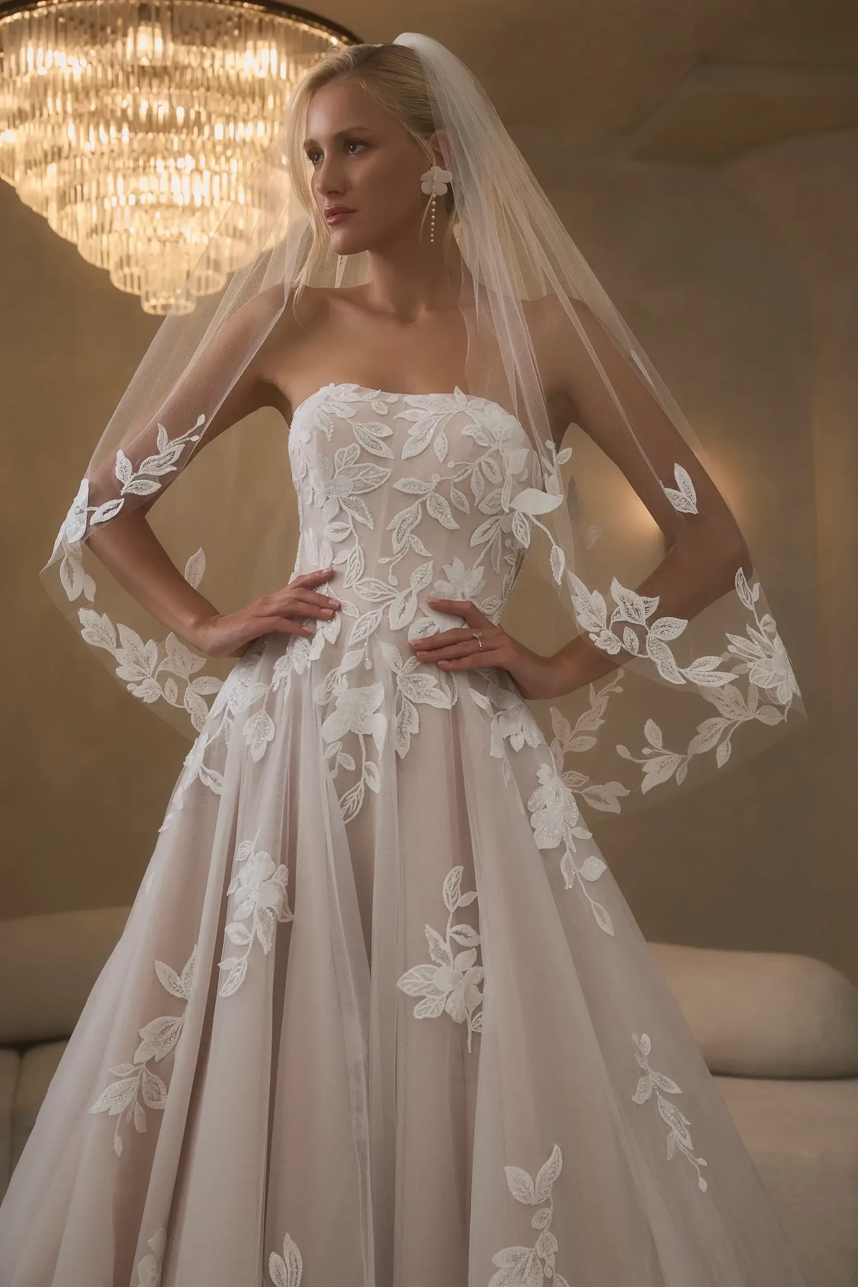 A bride wearing a strapless wedding gown adorned with floral lace details and a long veil, standing confidently in front of an elegant chandelier.
