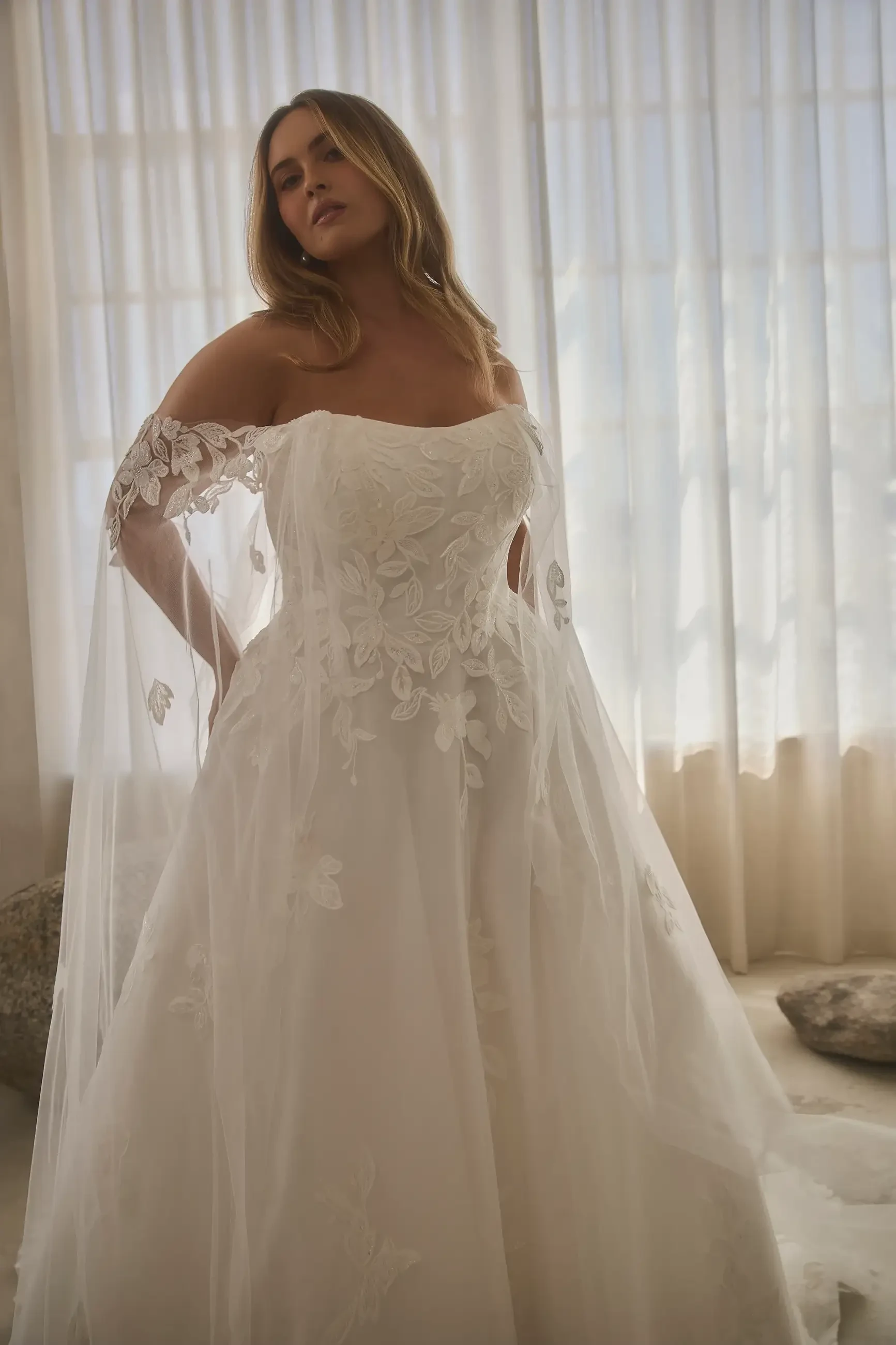A woman in an off-the-shoulder white wedding dress with floral details, standing gracefully in a softly lit room with sheer curtains.