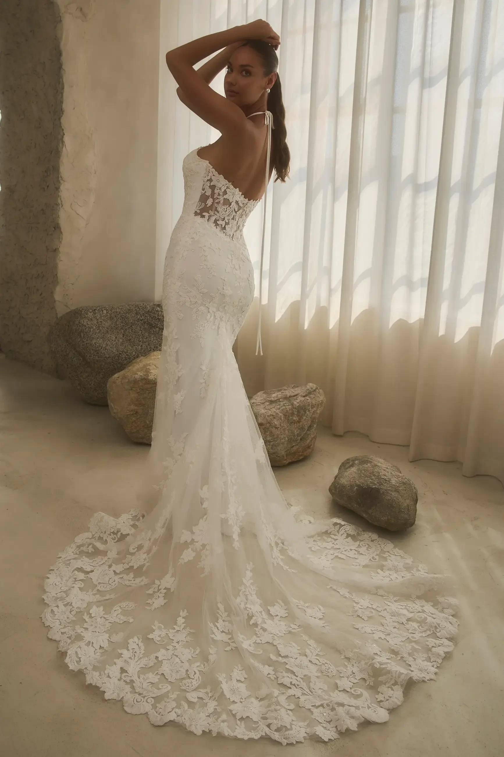 A woman in a white, lace wedding dress poses with her back to the camera, showcasing a long train, against a backdrop of soft lighting and natural stones.