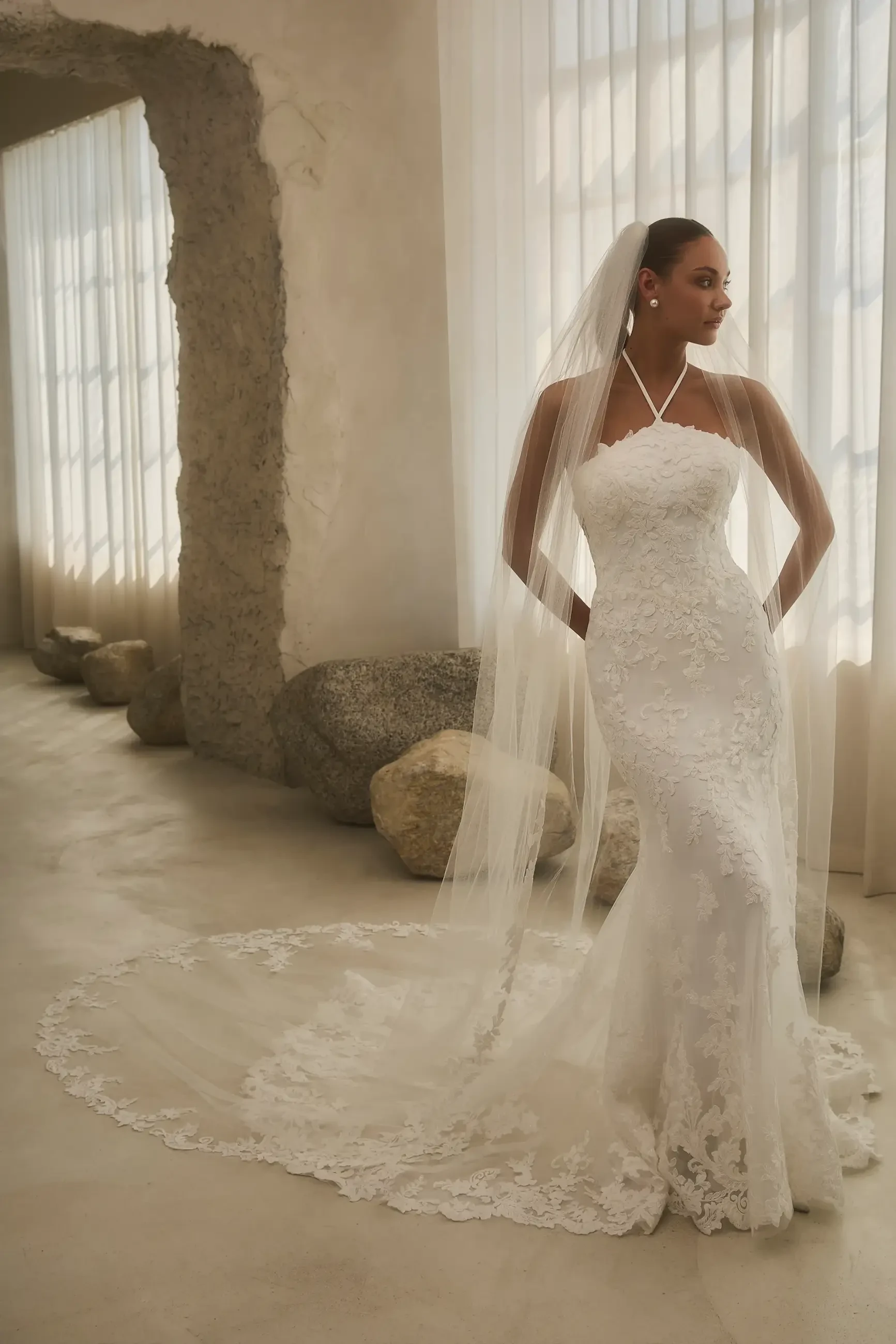A bride in a white wedding dress with lace details and a long veil, standing elegantly in a well-lit room with stone accents.