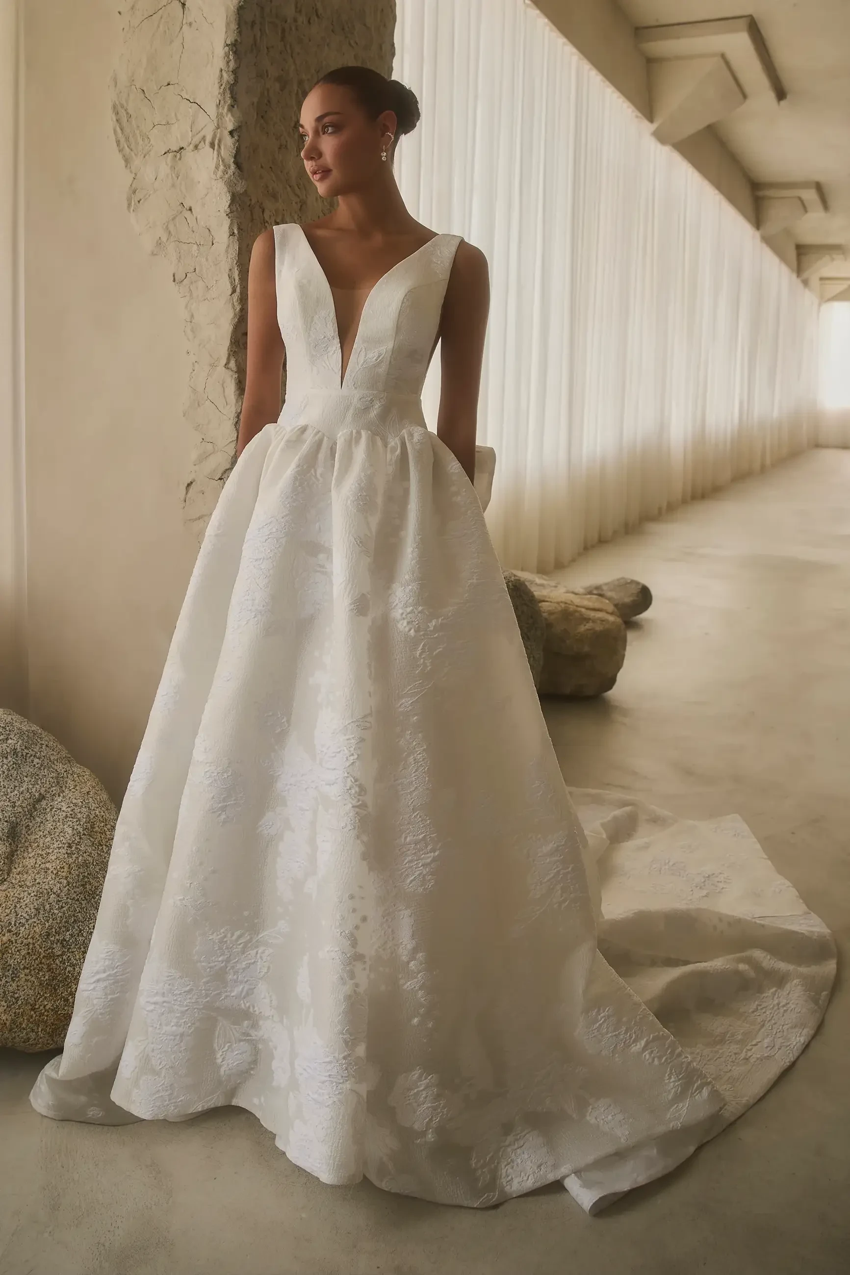 A woman wearing a white wedding dress with a deep V-neckline, standing in a well-lit, modern interior space.