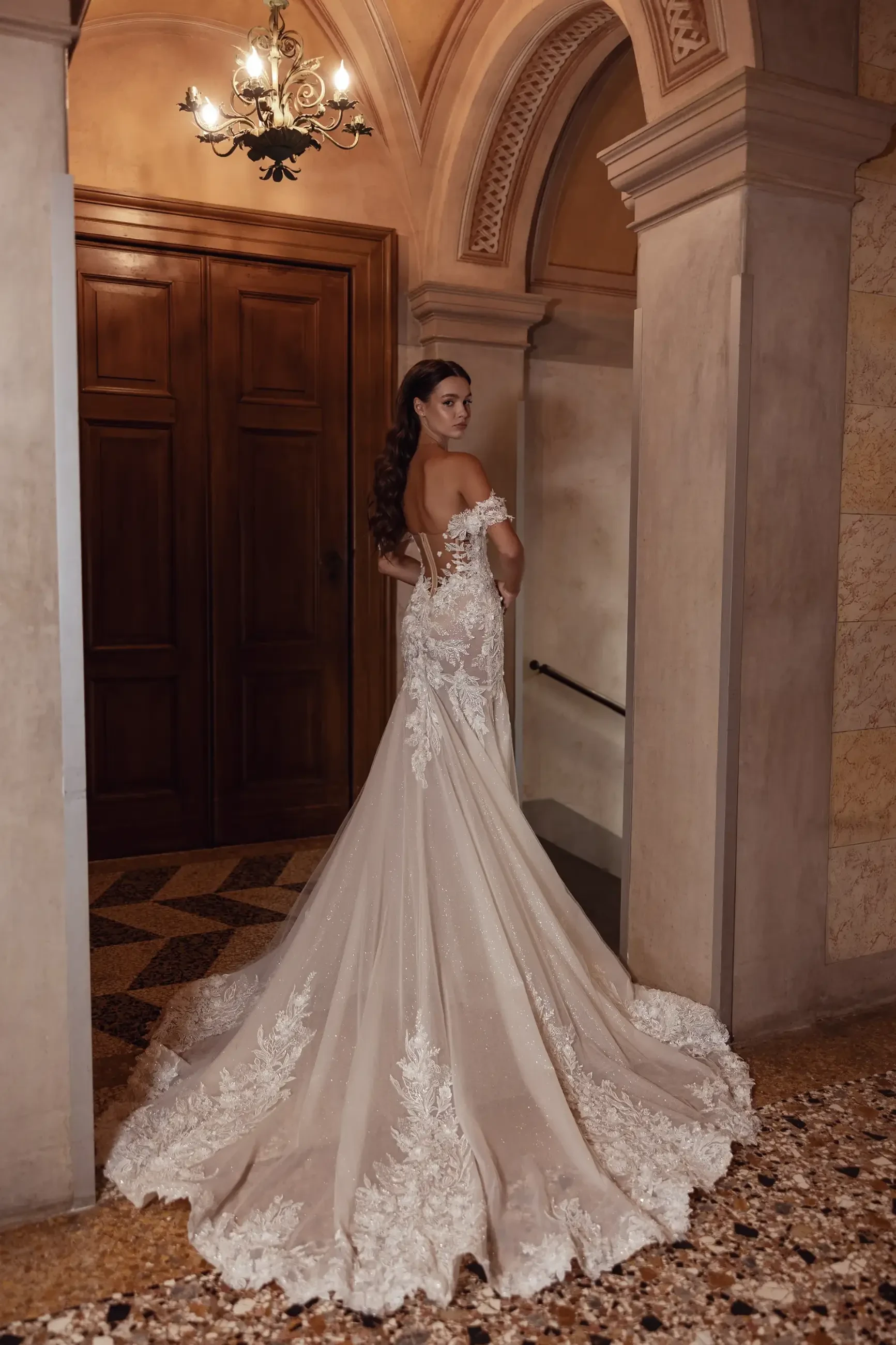 A bride in a lace wedding gown stands in a grand hallway with ornate arches and a chandelier. The dress features an elegant, flowing train.
