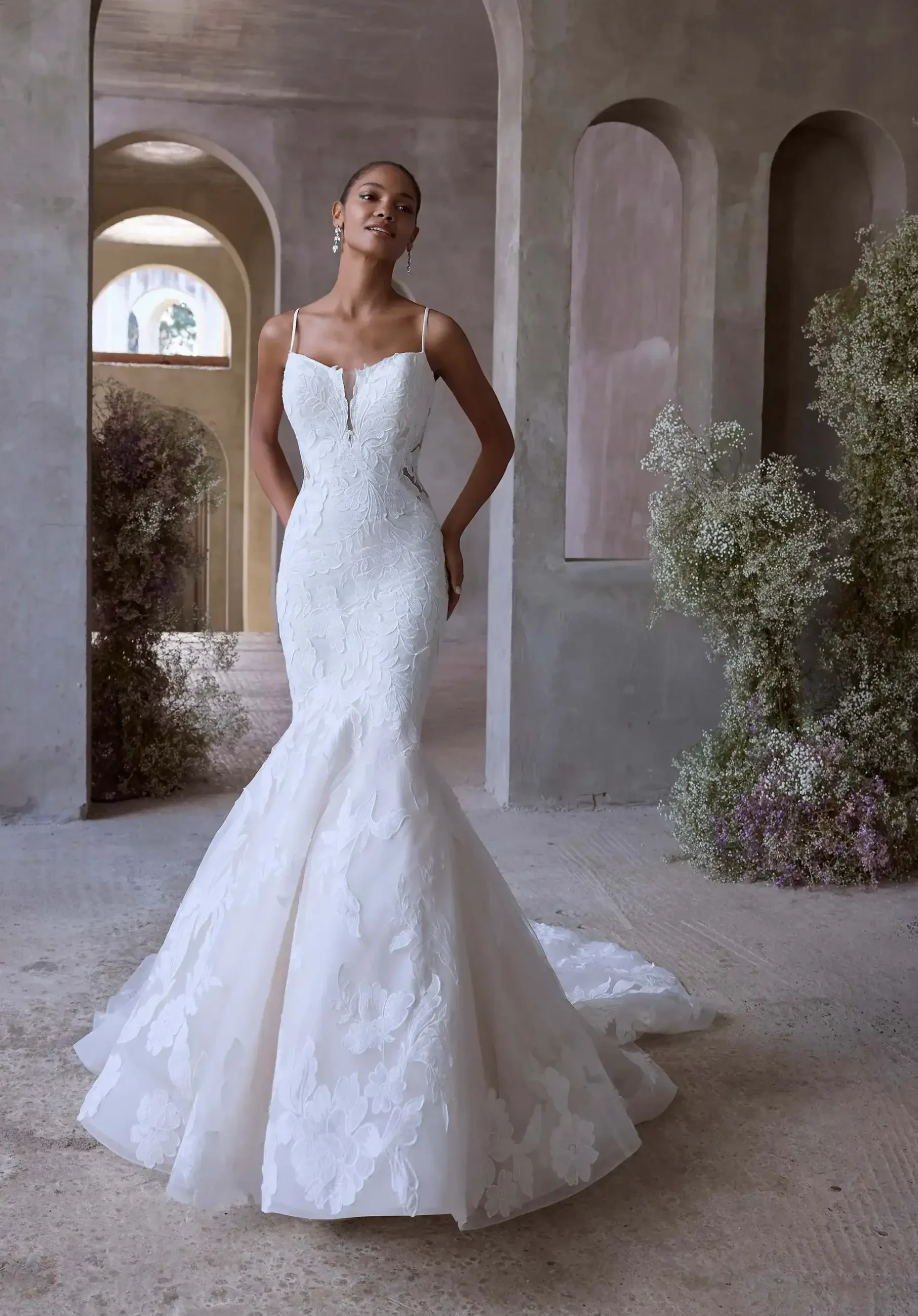 Model is wearing a mermaid wedding dress in soft tulle with a scoop neckline, sheer side cutouts, and floral lace appliqués, finished with a low back and flowing train.