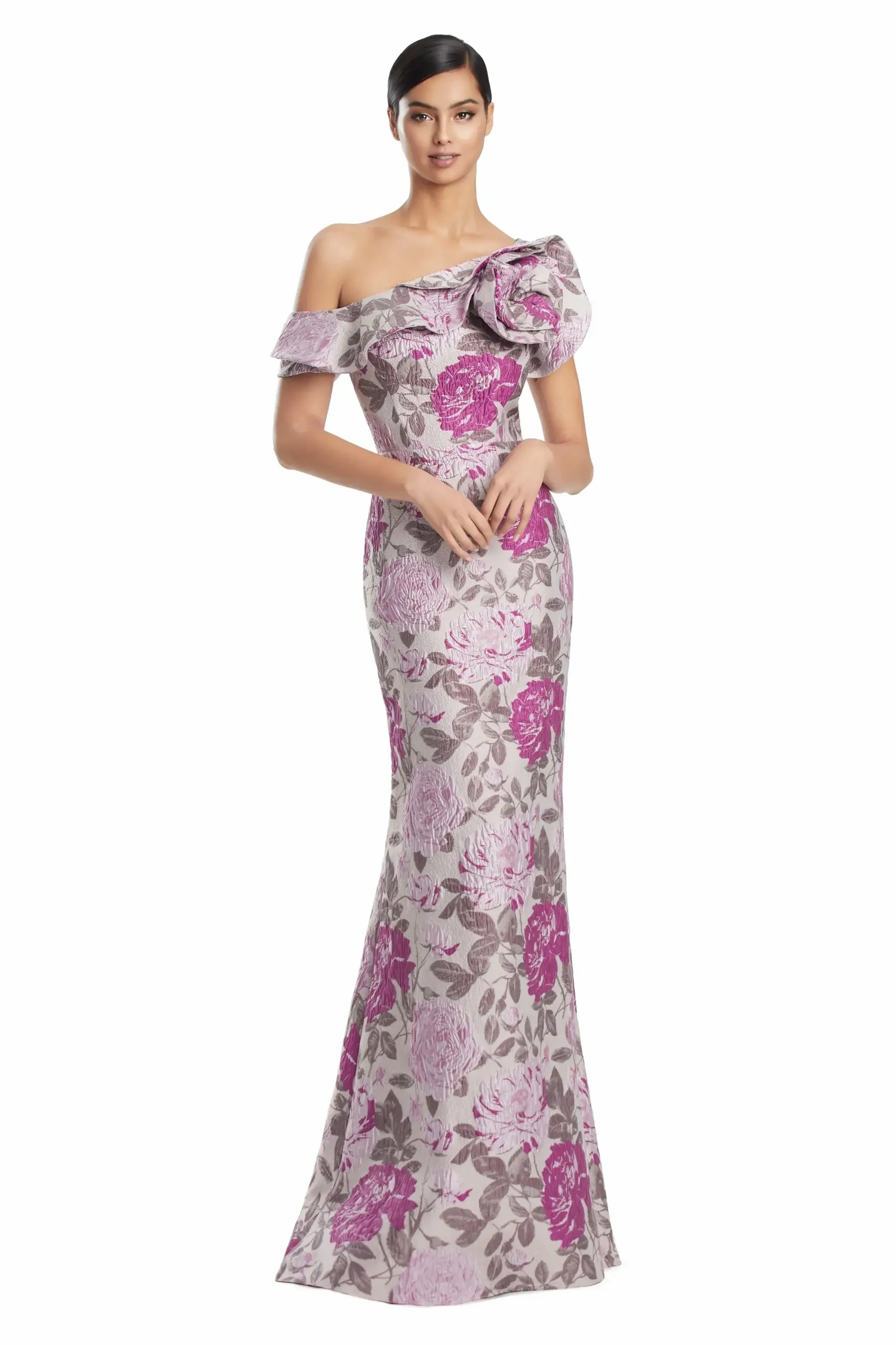 A model wearing a fitted, off-the-shoulder gown with a floral pattern in shades of pink and gray. The dress features a large ruffled detail on the shoulder and has a mermaid silhouette.