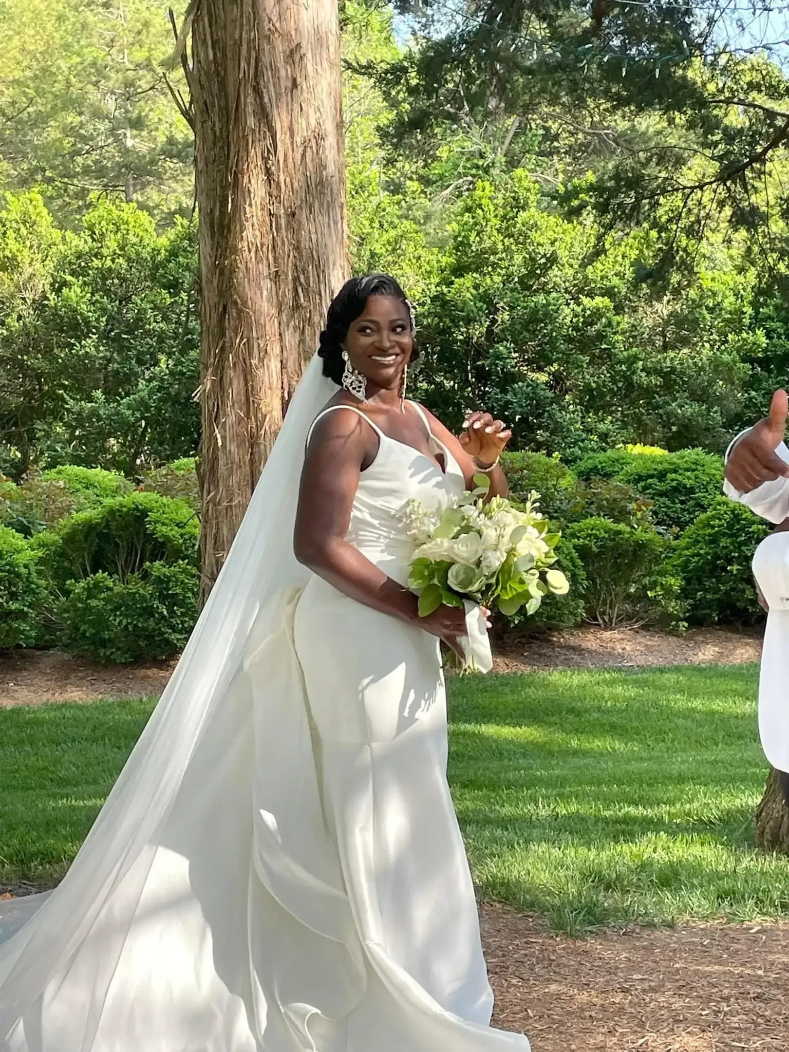 A woman in a white wedding dress holding a bouquet, smiling and waving. She is standing outdoors in a garden-like setting with greenery in the background.