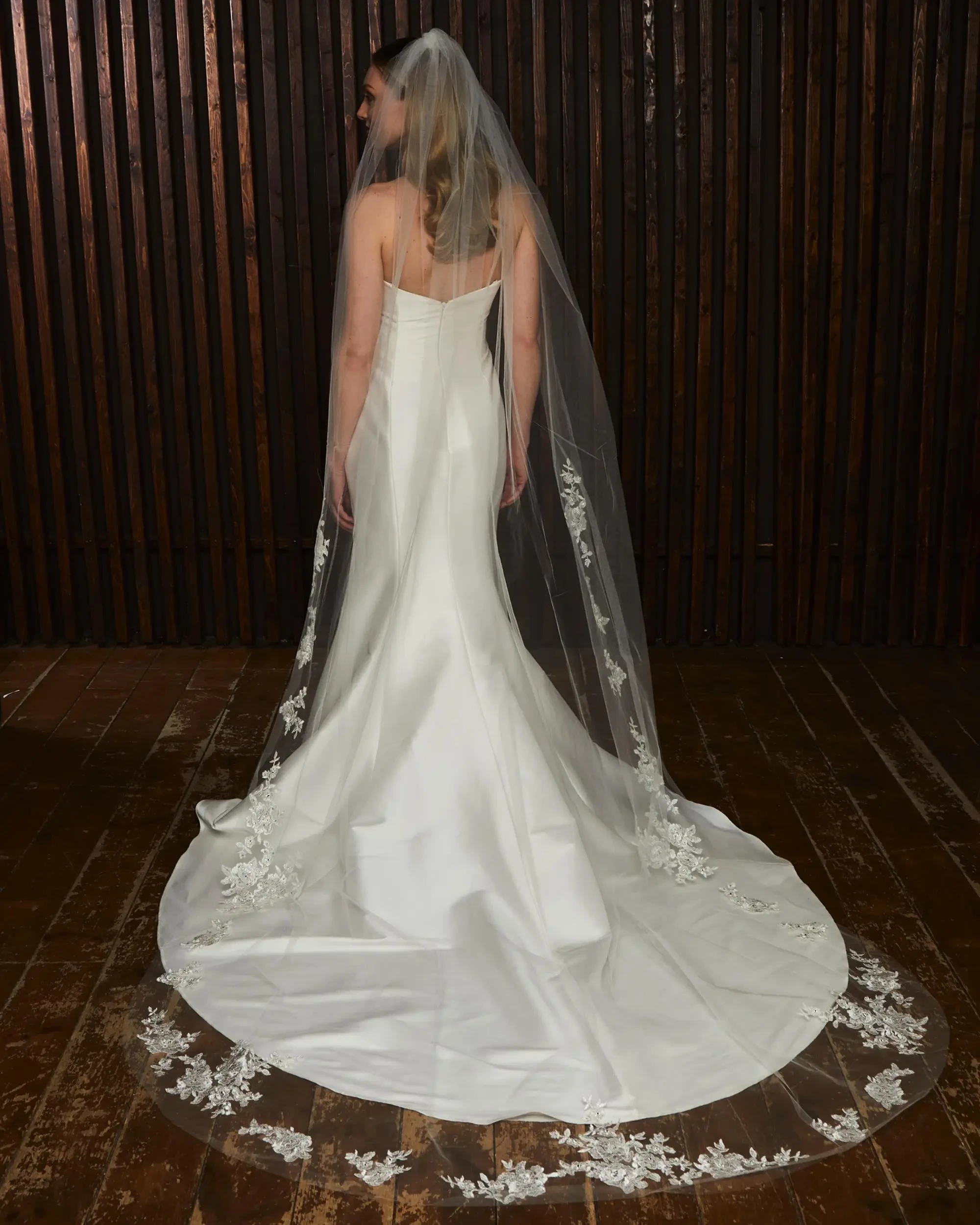 A woman in a wedding dress with a long veil, standing with her back to the camera. The dress has intricate floral details along the hem.