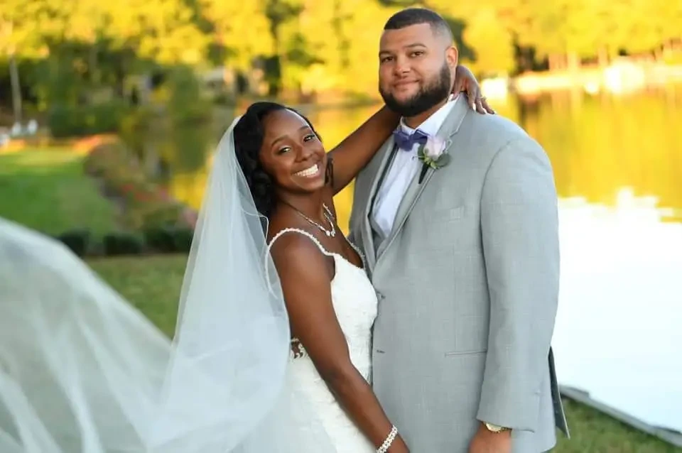 A happy couple posing for a portrait by a lake on their wedding day. The bride, wearing a white gown and veil, is smiling at the camera while the groom, dressed in a light gray suit with a bow tie, stands beside her.