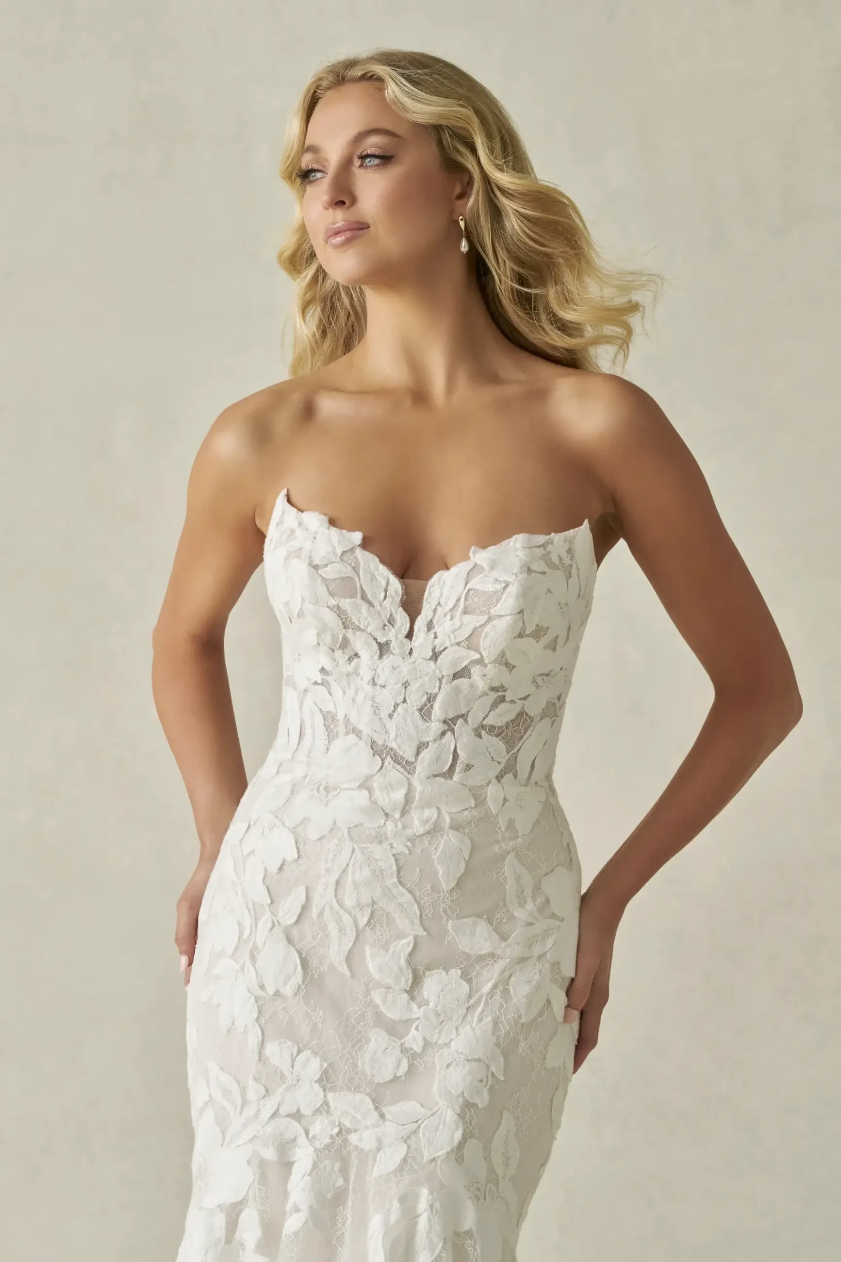 Blonde woman in strapless, floral lace, white wedding gown gazes thoughtfully to the side. The elegant, soft design conveys a sense of grace.