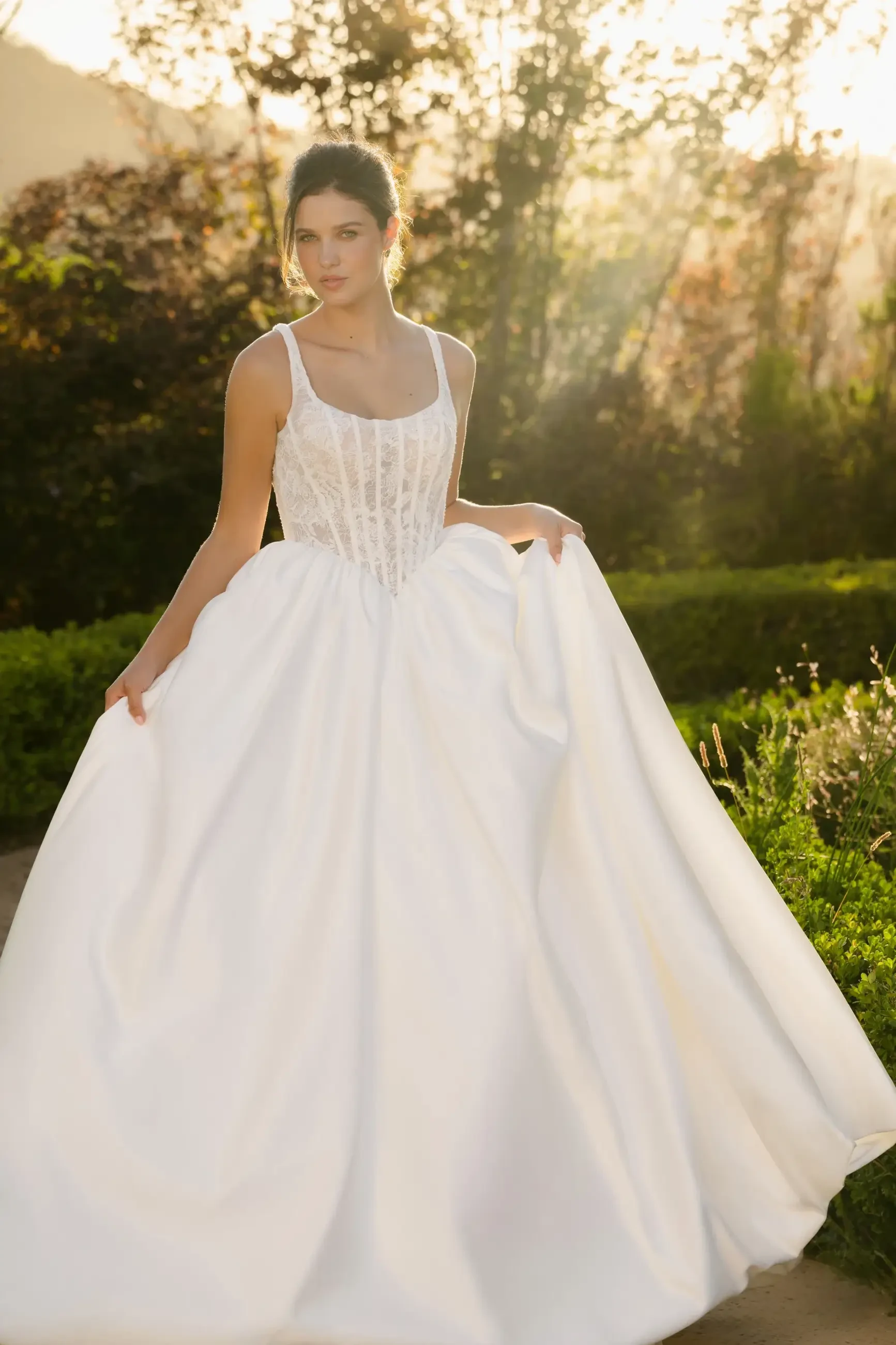 A woman in a flowing white dress stands in a garden, with sunlight filtering through the trees behind her.