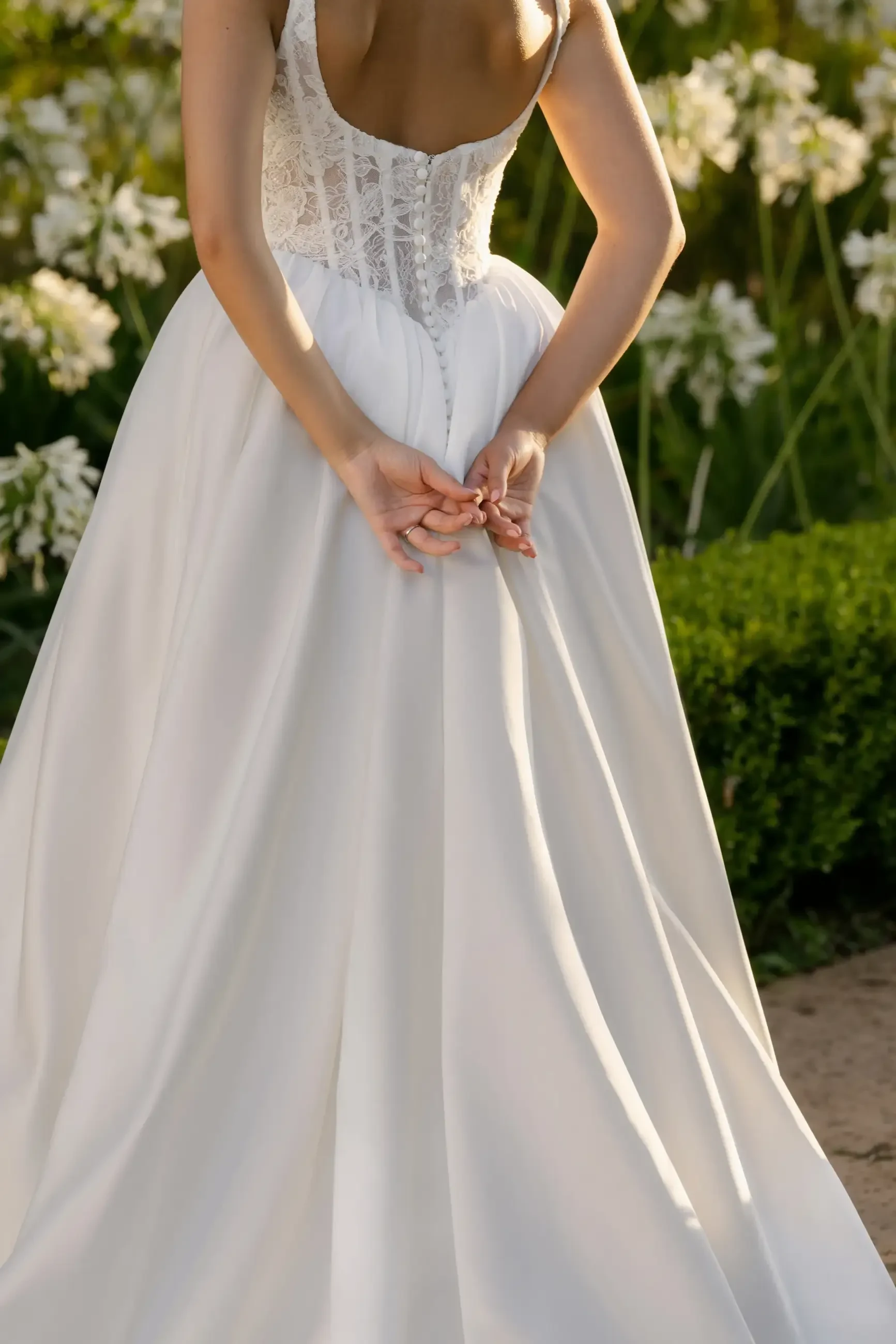 A woman in a white wedding dress holds the back of her gown in a garden with flowers.