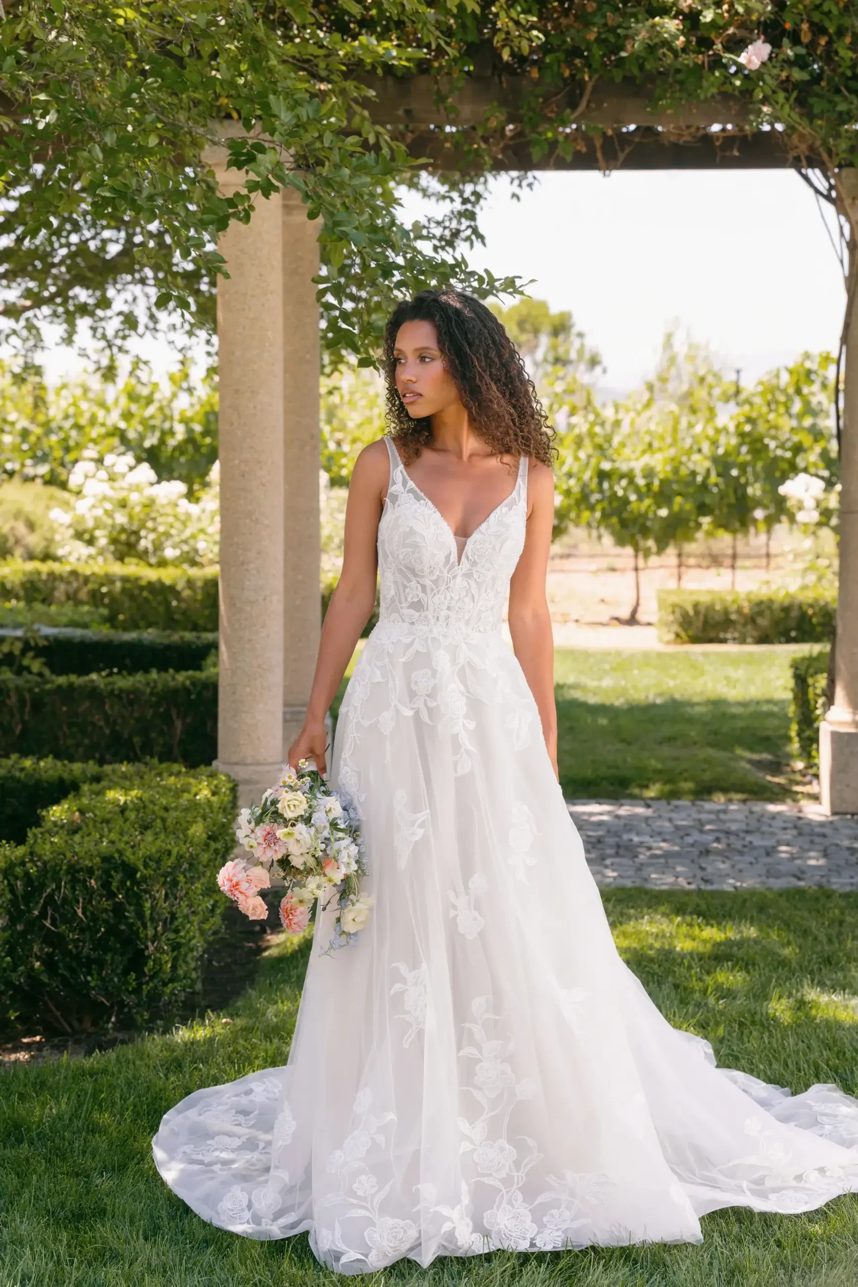 Bride stands outdoors in a white lace gown, holding a bouquet under a leafy pergola. The scene is serene and elegant, with lush greenery in the background.