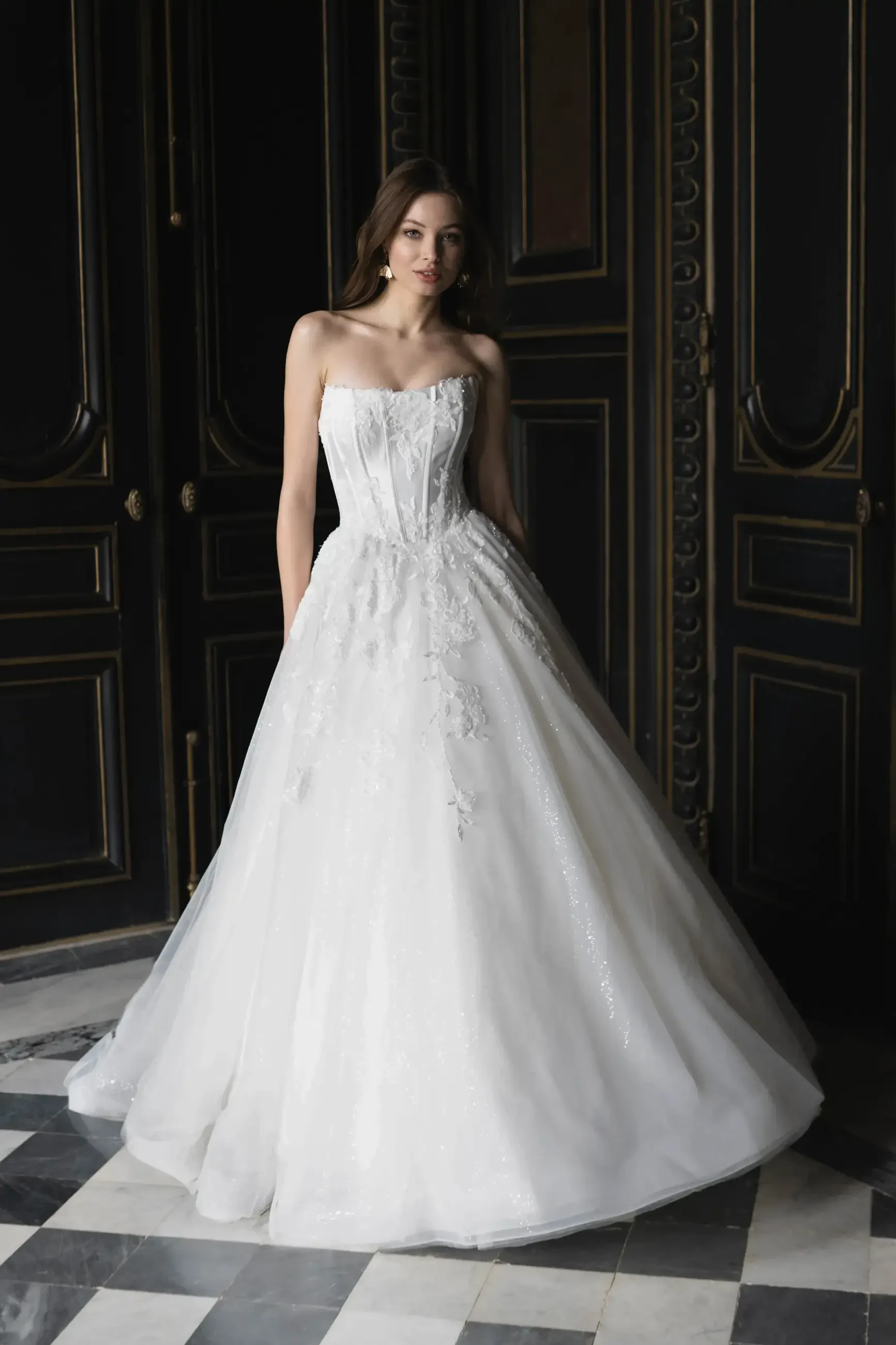 A model wearing a strapless white wedding dress stands gracefully in a dimly lit room with dark decor and patterned flooring.