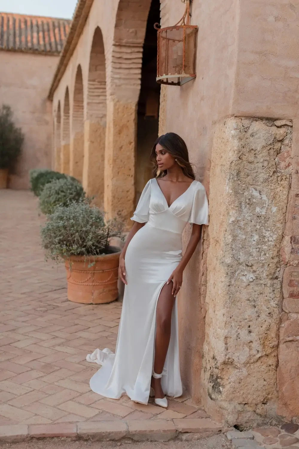 A model in a sleek white gown with a thigh-high slit poses against a rustic wall in an outdoor setting.