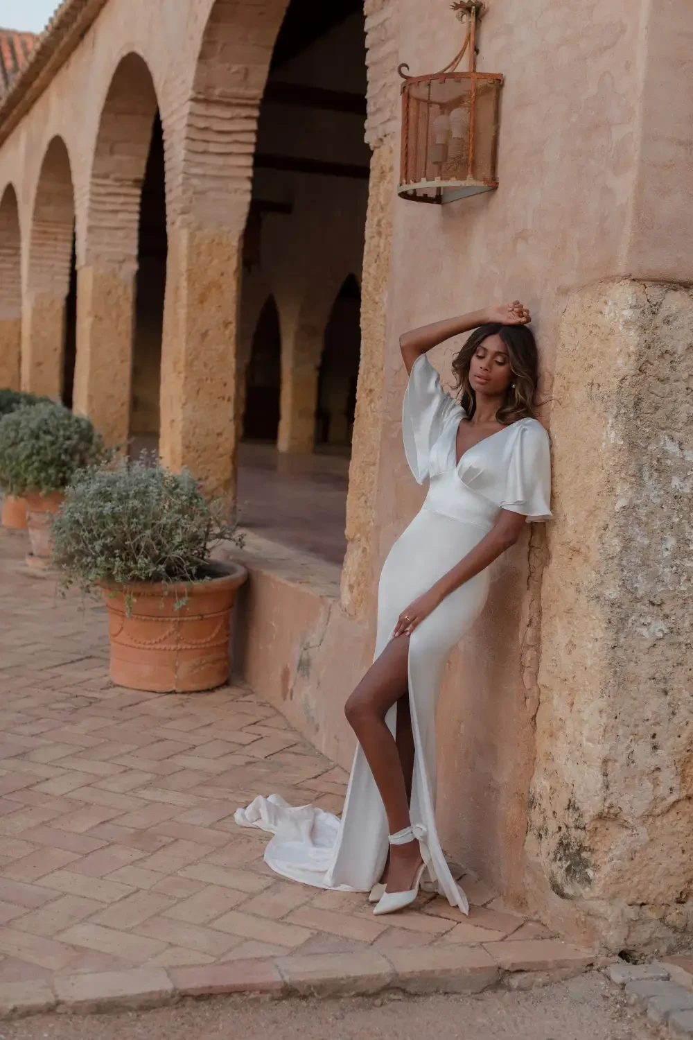 A model in a sleek white gown with a thigh-high slit poses against a rustic wall in an outdoor setting.