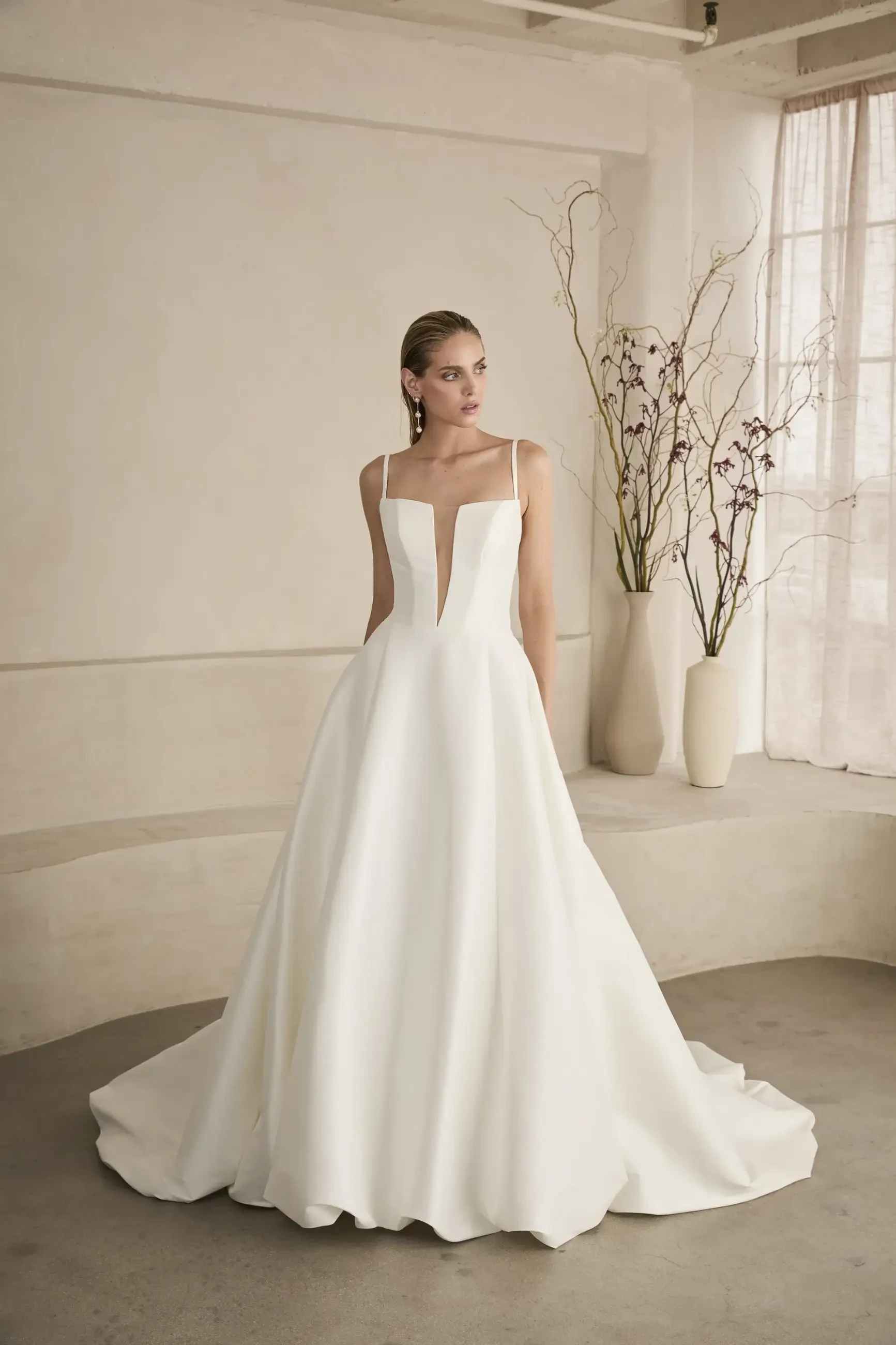 Model is wearing Dion, an A-line mikado gown with a plunging notched v-neckline, spaghetti straps, structured bodice with boning, and a sweeping 65-inch balloon-hem train.