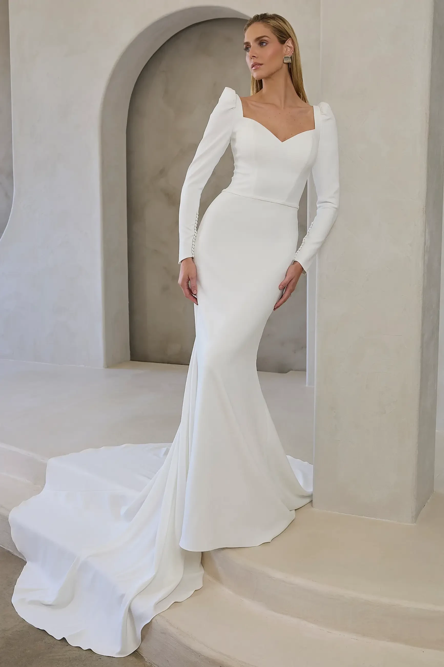Model is wearing Noa, a fit-and-flare gown with a sweetheart neckline, long gathered sleeves with covered buttons, a subtle v-back, and a dramatic 76-inch train.