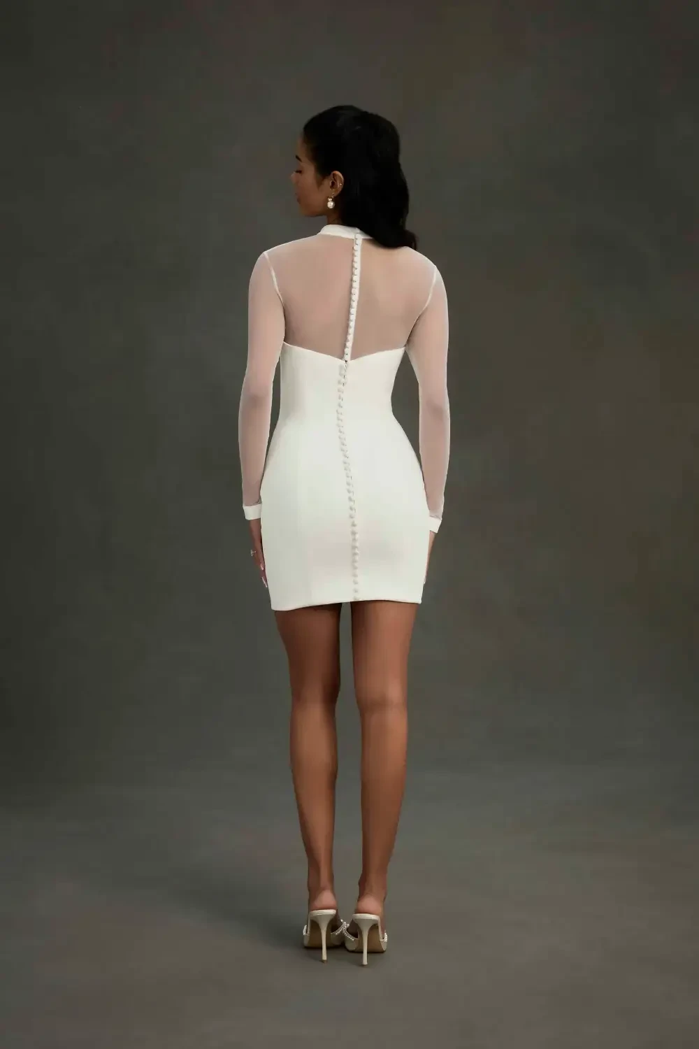 A woman stands confidently in a fitted white dress with sheer long sleeves, against a dark gray background.